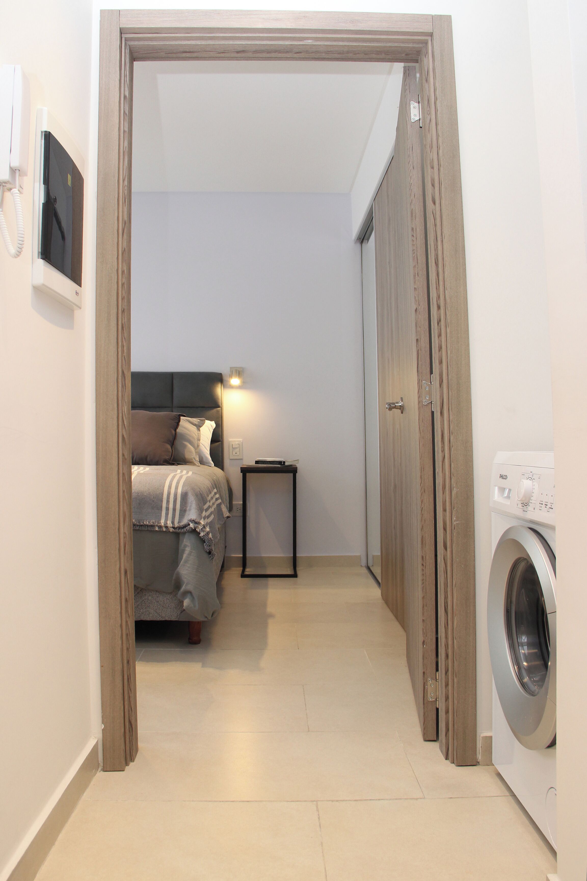 A narrow hallway leading to a bedroom with a bed, bedside table, and a washing machine.