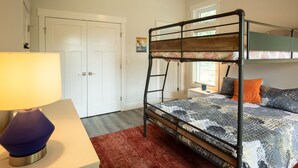 A bunk bed with a patterned bedspread, a desk with a lamp, and a red rug in a room with white walls and a door. - Modern Soundfront Studio, Pier access, Sunsets, Fishing, Watersports, Beach (Salvo)