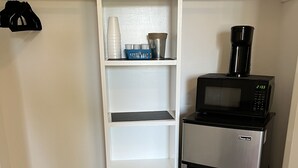 A compact kitchen with a stainless steel refrigerator, a black microwave, and a coffee maker on top. There is a white shelving unit with cups and a basket. A black trash bin is on the floor. - Modern Soundfront Studio, Pier access, Sunsets, Fishing, Watersports, Beach (Salvo)