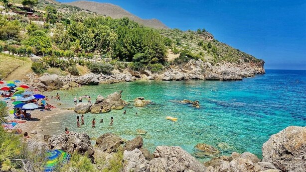 A coastal beach with clear turquoise water, rocky shore, and umbrellas.