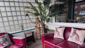 Een rode leren bank met luipaardprint kussens, een klein houten bijzettafeltje en een grote kamerplant in een kamer met een raam met patroon en uitzicht op gebouwen.