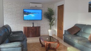 A living room with a TV on the wall, a wooden cabinet, a glass coffee table, two leather sofas, and a potted plant.