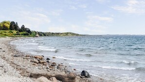 Een strand met kiezels en rotsen, kalm water en een heuvellandschap op de achtergrond.