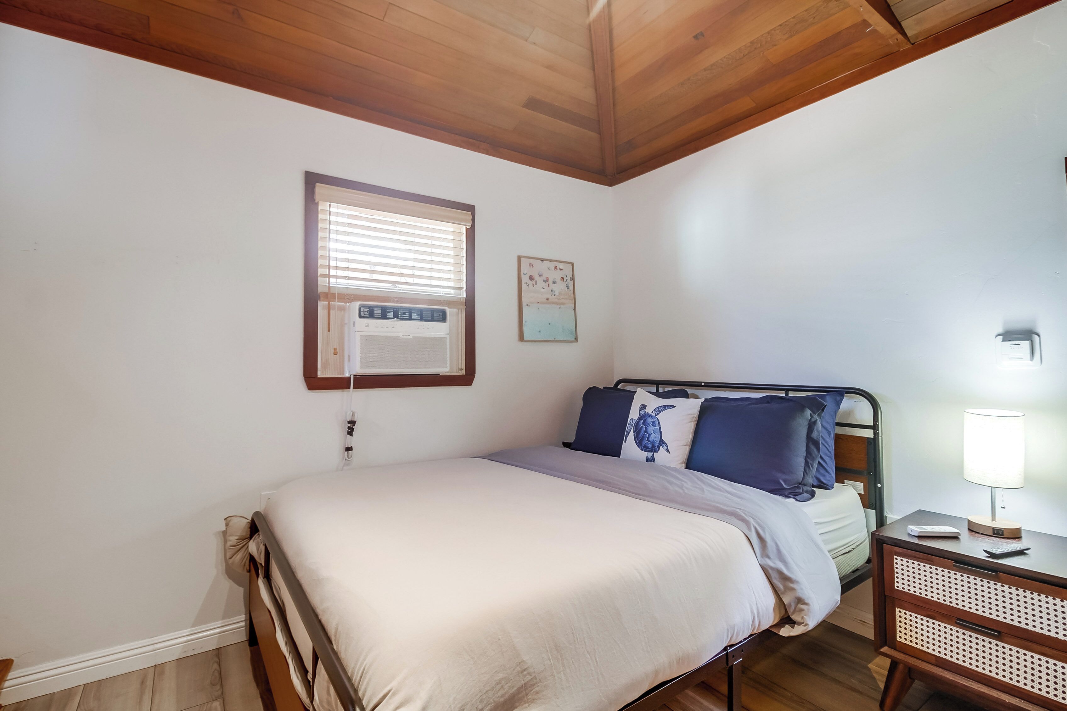 Un dormitorio con cama, una mesita de noche con lámpara, una ventana con persianas y un aire acondicionado de pared.