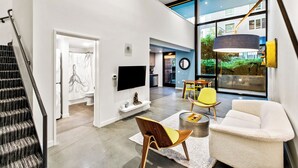 A modern living room with a staircase, a flat-screen TV, a dining area with yellow chairs, and large windows offering a view of the outdoors. - Posh Lofty Pad in Central Redmond (Redmond)