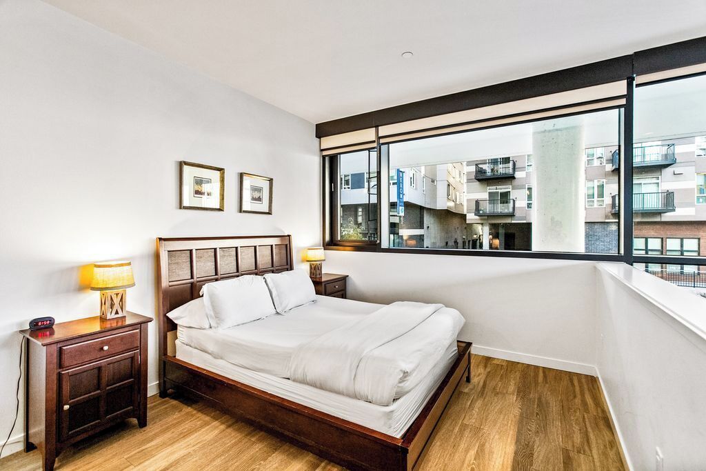 Een moderne slaapkamer met een groot bed, een houten hoofdeinde, twee nachtkastjes met lampen en een uitzicht op gebouwen door een groot raam.