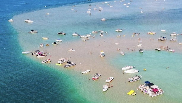 An aerial view of a beach with numerous boats anchored near the shore and people enjoying the beach. - Torch Lake Sandbar Escape (Rapid City)