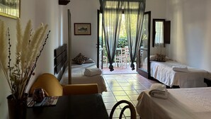 Ein Hotelzimmer mit zwei Betten, einer Essecke inklusive Tisch und Stühlen, einer Vase mit Trockenpflanzen und Blick auf den Garten durch durchscheinende Vorhänge.