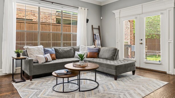 Ein Wohnzimmer mit einem grauen Ecksofa, zwei runden Tischen, einem Teppich mit Fischgrätmuster und Blick auf eine Ziegelwand durch große Fenster.