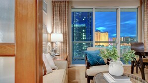 A modern hotel room with a large window offering a city view, a sofa, a coffee table, and a dining area with a chair. - The Suite Spot@MGM Signature Balcony Strip View (Las Vegas)