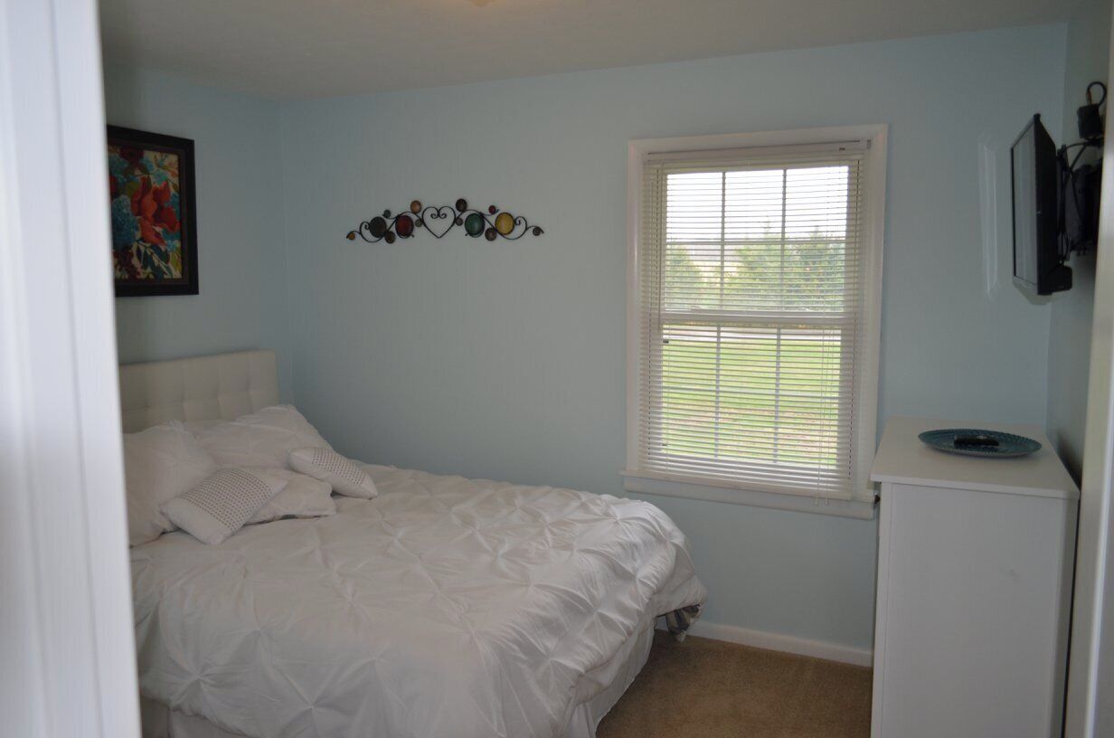A bedroom with a bed, a window with blinds, a small white cabinet, and a wall decoration.