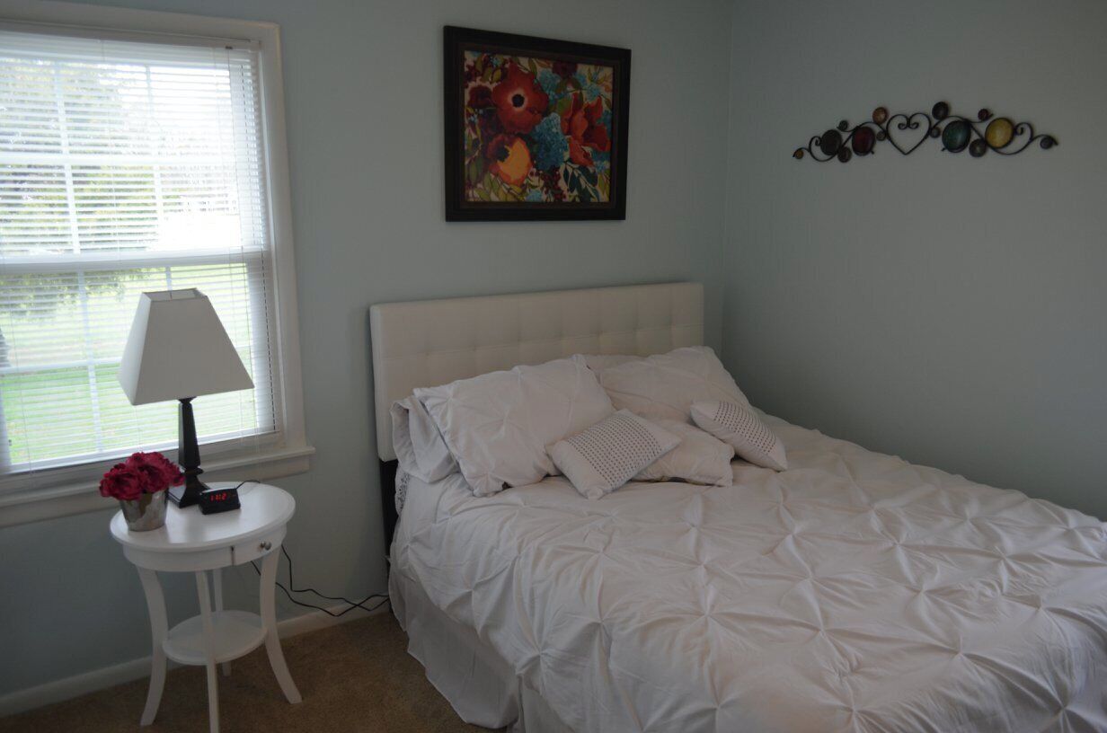 A bedroom with a white bed, a bedside table with a lamp, and a floral painting on the wall.