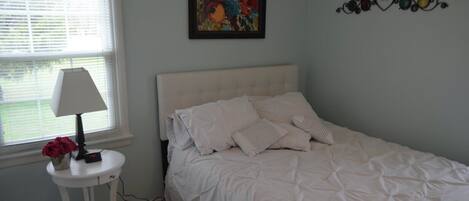 A bedroom with a white bed, a bedside table with a lamp, and a floral painting on the wall.