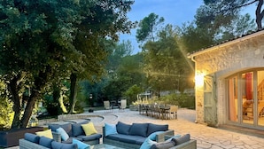 Un patio al aire libre con mobiliario de mimbre, piso de piedra y un edificio de piedra con ventanas grandes.