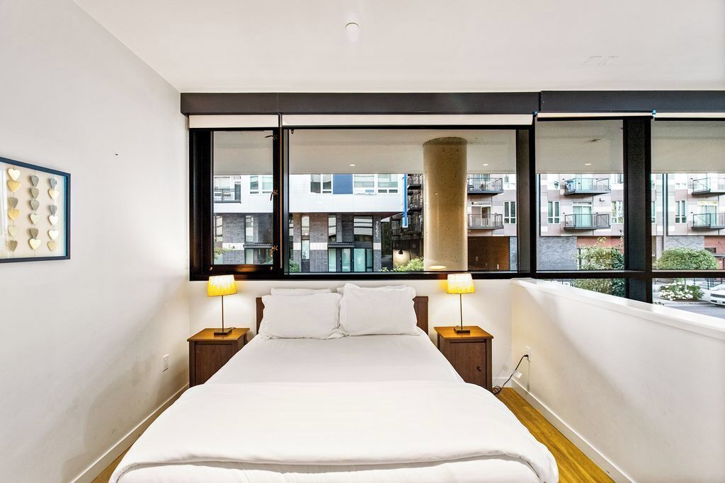 A modern hotel room with a large bed, two bedside tables, a window with a view of other buildings, and a wall-mounted light fixture.