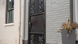A metal door with decorative patterns, a white brick wall, and a planter with dried plants.