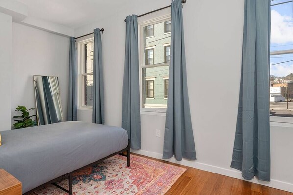 A bedroom with a bed, a window with curtains, a mirror, and a patterned rug.