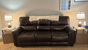 A brown leather sofa with a white throw blanket, flanked by two bedside tables with lamps, a framed 'home' sign, and a window with a view. - 1-800 We got the stay (Amarillo)