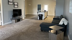 A spacious living room with a dark-colored sofa, a small wooden table, and a TV on a stand. There is a kitchen area with a sink and a countertop visible in the background. - 1-800 We got the stay (Amarillo)