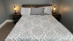 A bedroom with a patterned bedspread, a wooden headboard, two bedside tables with lamps, and a carpeted floor. - 1-800 We got the stay (Amarillo)