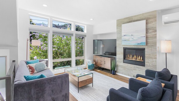 A modern living room with a sofa, armchairs, a glass coffee table, a fireplace, a TV, and large windows with a garden view. - Golden Gardens Getaway Beach Waterfront (Seattle)