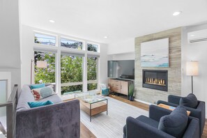 A modern living room with a sofa, armchairs, a glass coffee table, a fireplace, a TV, and large windows with a garden view. - Golden Gardens Getaway Beach Waterfront (Seattle)
