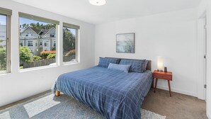 A bedroom with a large bed, a nightstand with a lamp, a framed picture on the wall, and a view of a house through the window. - Golden Gardens Getaway Beach Waterfront (Seattle)