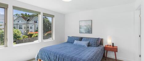 A bedroom with a large bed, a nightstand with a lamp, a framed picture on the wall, and a view of a house through the window.