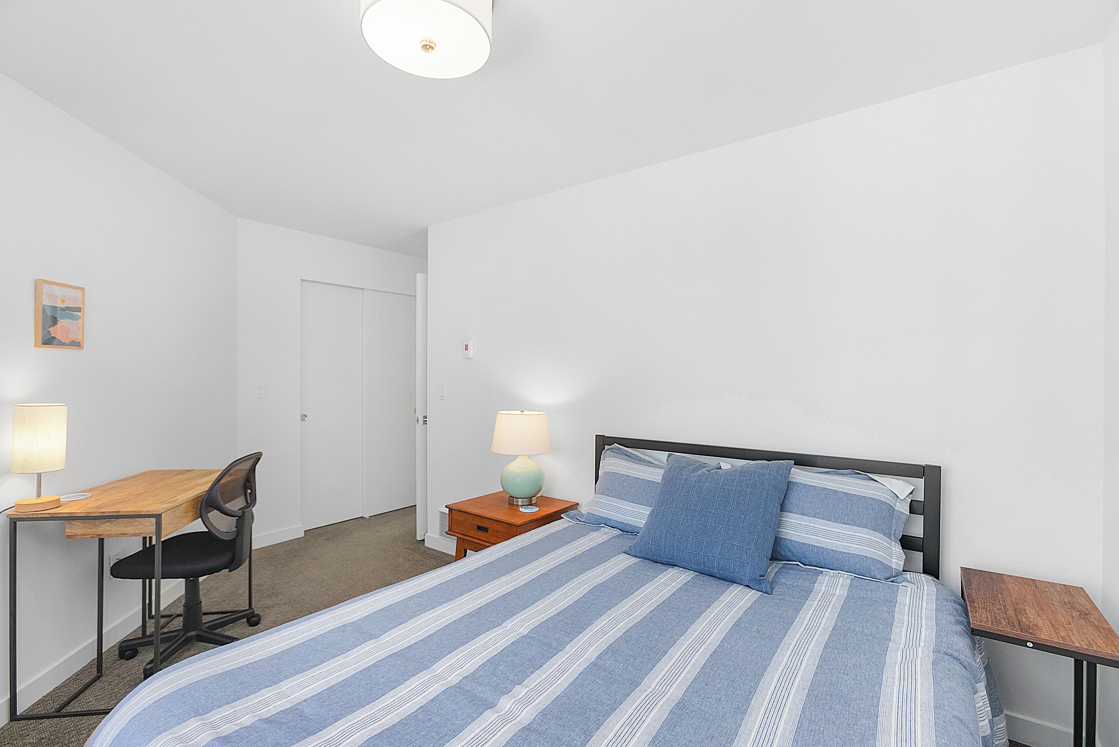 A bedroom with a bed, a desk, a chair, a nightstand, and a lamp.
