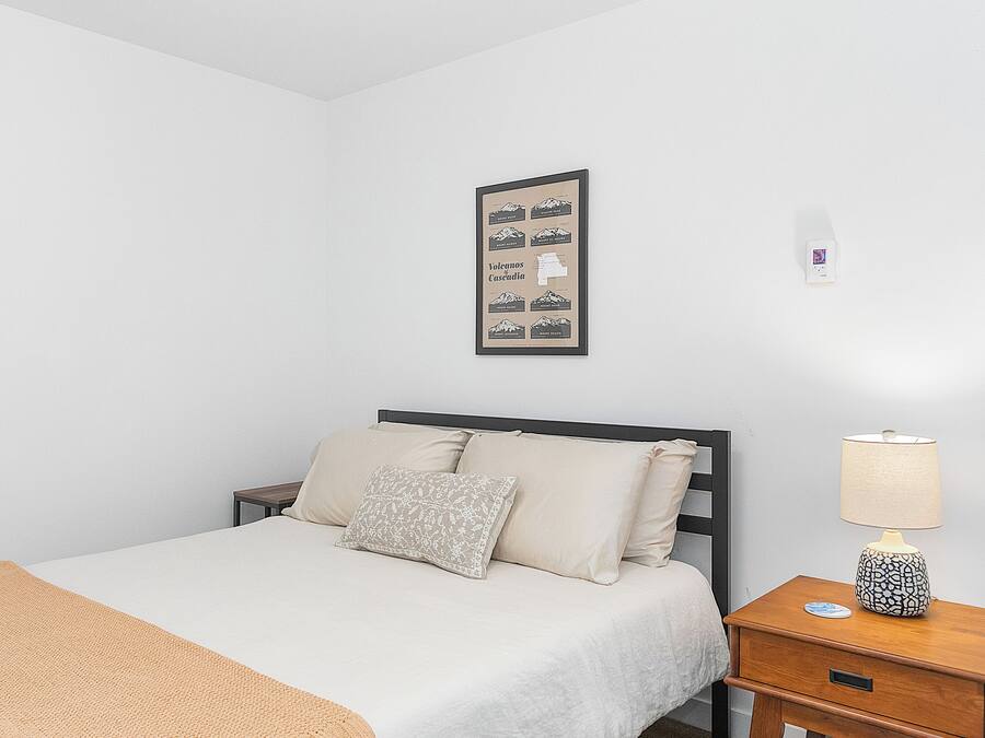 A neatly made bed with a beige blanket and pillows, a bedside table with a lamp, and a framed map on the wall.