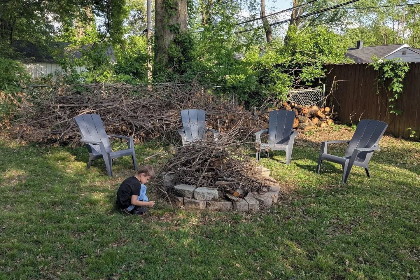 A backyard with a pile of branches, three Adirondack chairs, and a child playing.