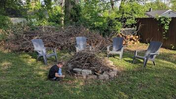 A backyard with a pile of branches, three Adirondack chairs, and a child playing.