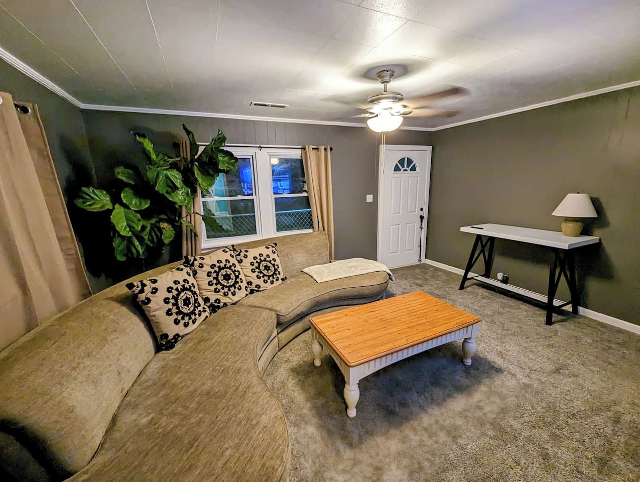 A living room with a sofa, a coffee table, a ceiling fan, a window with curtains, and a door.