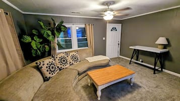 A living room with a sofa, a coffee table, a ceiling fan, a window with curtains, and a door.
