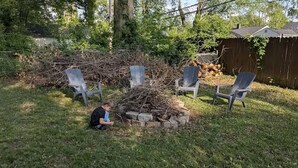 Patio trasero con un brasero rodeado por cuatro sillas Adirondack, una valla de madera y una pila de leña.