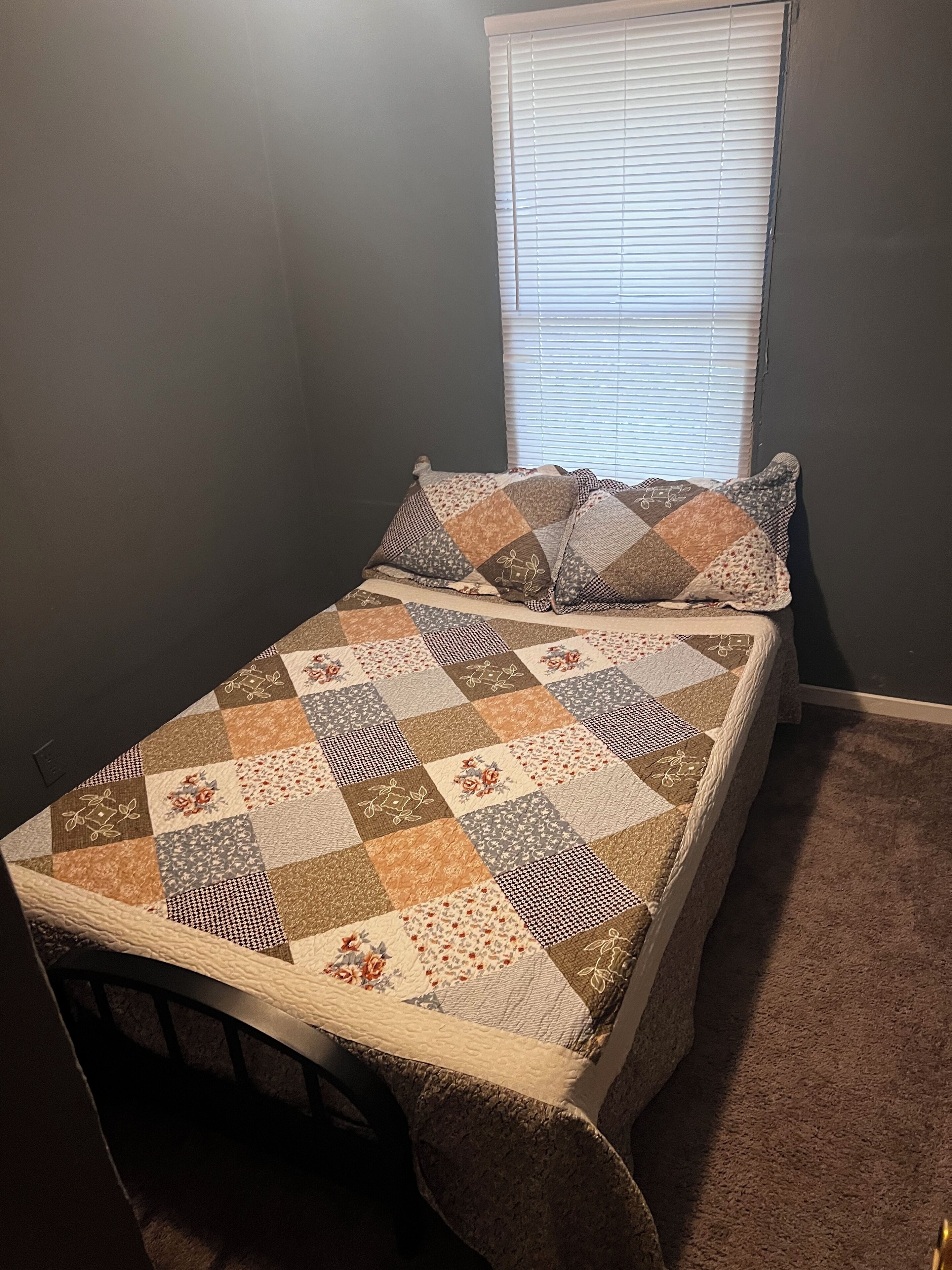 Een klein bed met een sprei, een raam met zonwering en een donkere muur.