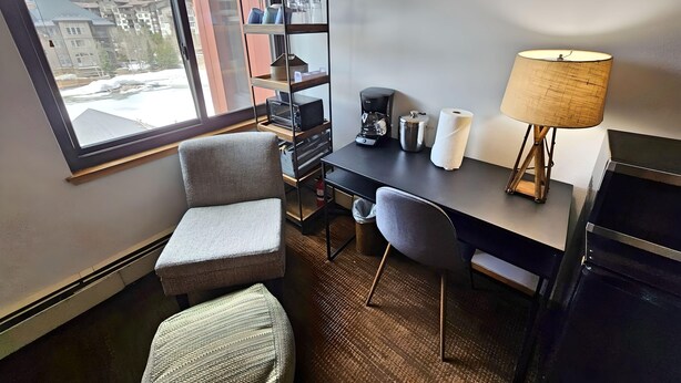 A hotel room with a desk, two chairs, a lamp, a coffee maker, a shelf with items, and a view of buildings and mountains through the window. - Village square 442, Center Village Copper Mountain,Pet Friendly includes parking (Copper Mountain)
