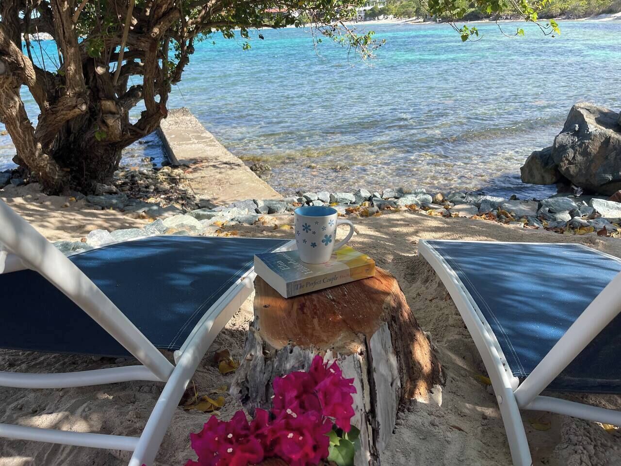 Un espai relaxant a la vora de la platja amb dues gandules, una taula de fusta, un llibre i una tassa, i un arranjament floral.
