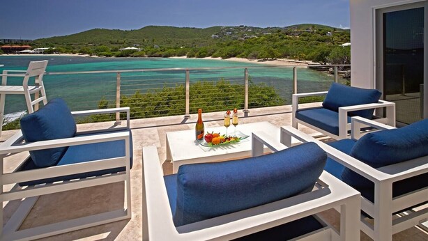 Un balcon agrémenté de meubles blancs et d’une table où sont disposés des boissons et des fruits, offrant une vue sur une plage et des eaux turquoise.