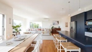 A modern kitchen with a central island, white bar stools, and a dining area with a long table. - Salty Ocean View Escape - Torquay (Surf Coast)