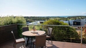 A wooden deck with a round table and four chairs, overlooking a landscape with greenery and buildings. - Salty Ocean View Escape - Torquay (Surf Coast)