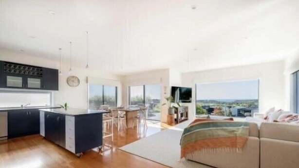 A modern kitchen with a central island, a dining area with chairs, a sofa, and a large window offering a view. - Salty Ocean View Escape - Torquay (Surf Coast)