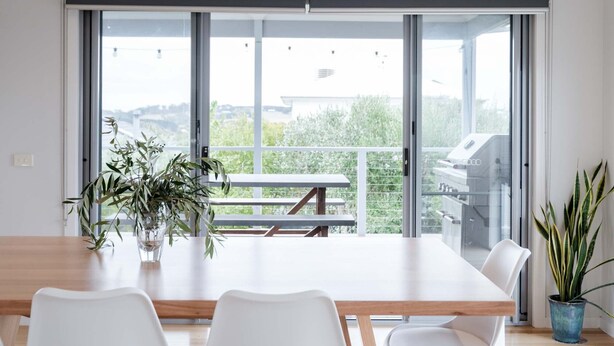 A modern dining area with a large table, white chairs, and a view of a balcony with a grill. - Torquay Beach Holiday Homebase (Surf Coast)
