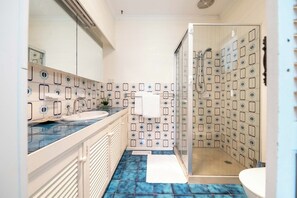 A bathroom with a glass-enclosed shower, a sink with a mirror, and patterned tiles on the walls and floor. - Country Seaside Escape - Torquay (Surf Coast)