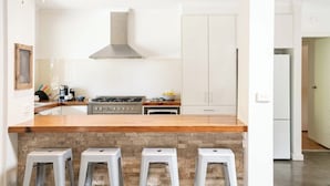 A modern kitchen with a central island, a gas stove, and a stainless steel range hood. There are three bar stools in front of the island. - Country Seaside Escape - Torquay (Surf Coast)