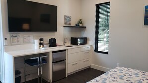 Une chambre d’hôtel compacte dotée d’une kitchenette, d’un lit avec une couette à motifs bleus et blancs, d’une grande fenêtre avec stores, d’un téléviseur à écran plat fixé au mur et d’une petite étagère ornée d’une plante et d’une étoile de mer décorative.