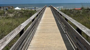 Une promenade en bois mène à une plage où l’on aperçoit, au loin, des tentes et des personnes se prélassant sur le sable.
