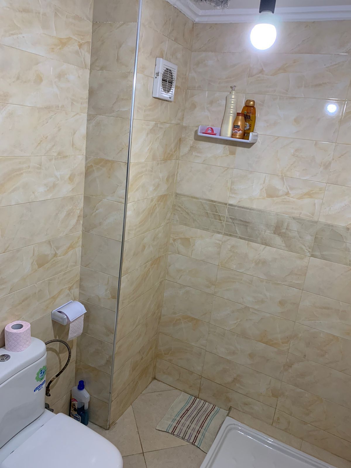 Een badkamer met een toilet, douche en wastafel. Er is een plank met toiletartikelen en een rol wc-papier.