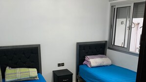 A bedroom with two beds, a small bedside table, and a window with blinds. - Spacious 3-bedrooms apartment (Tétouan)
