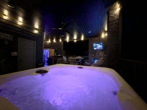 A hot tub with a ceiling fan, a TV screen, and a seating area with chairs and a table.